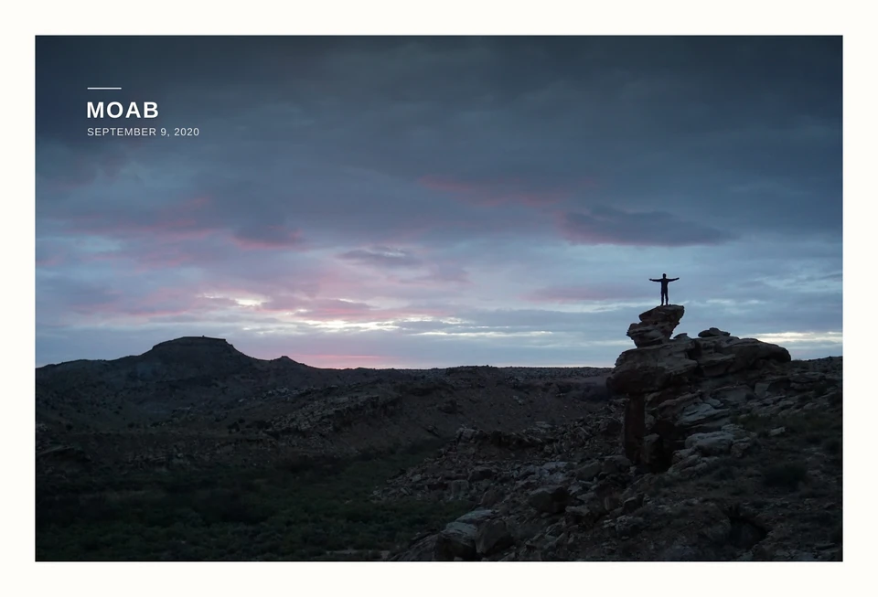 A photograph of a person standing with arms outstretched on a desert rock beneath a pink and blue sky. Moab, September 9, 2020.
